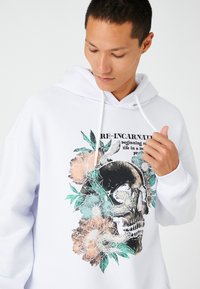 Biała bluza z kapturem z grafiką czaszki otoczoną kolorowymi kwiatami i liśćmi. Napis brzmi "RE-INKARNACJA". Miękki materiał, casualowy krój.