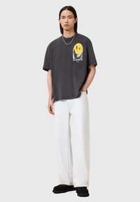 Mørkegrå oversized t-shirt med et gult smiley grafik og "ALLSAINTS" logo. Kombineret med hvide bukser med brede ben og sorte sko.
