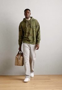 Olivovo zelená mikina s vzorom listov, svetlé džínsy, biele tenisky a béžová taška cez plece s tmavými popruhmi. Jednoduchý, neformálny outfit.