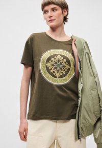 Ελιάς πράσινη βαμβακερή μπλούζα με στρογγυλή λαιμόκοψη, διακοσμημένη με ένα κεντρικό χρυσό και πράσινο λογότυπο. Ελαφρύ ύφασμα, κοντά μανίκια, άνετη εφαρμογή.