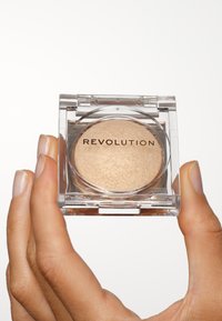 Revolution highlighter i en klar, kvadratisk emballasje med en glatt, skimrende gulloverflate. Hender som holder emballasjen.