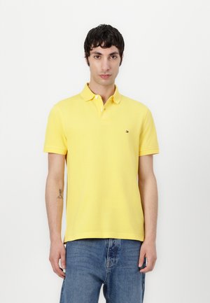 Gelbes Polohemd aus Baumwolle, mit Kragen, kurzen Ärmeln und einem kleinen Logo auf der linken Brust. Getragen mit blauen Jeans.