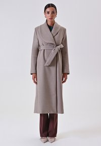 Cappotto di lana beige con grande colletto ripiegato, una cintura in vita e due bottoni. Indossato sopra pantaloni marroni e scarpe chiare.