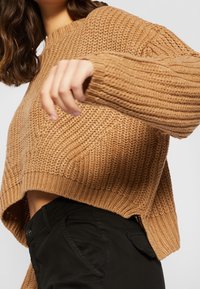 Femme portant un pull ample en tricot de couleur beige avec un ourlet plus long à l'arrière et un pantalon noir, levant son bras gauche partiellement devant son corps.