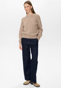 Svetlo rjav, teksturiran pulover s okroglim ovratnikom, v kombinaciji s temno modrimi širokimi jeans hlačami. Model nosi bež gležnjarje.