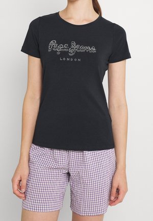 Frau trägt ein schwarzes T-Shirt mit kurzen Ärmeln und dem glitzernden "Pepe Jeans London"-Logo sowie lila karierte Shorts vor einem schlichten Hintergrund.