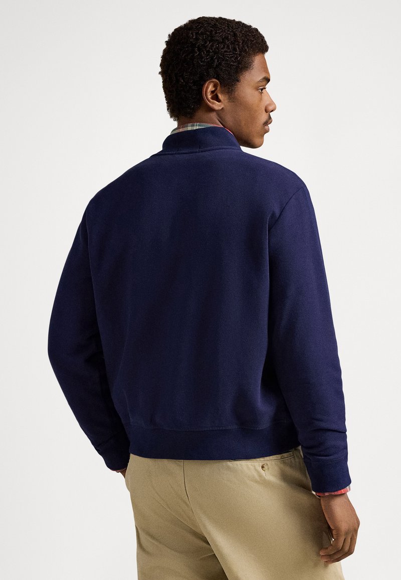 Navy sweatshirt with a stand-up collar, ribbed cuffs, and hem. Smooth fabric with a relaxed fit, worn over a plaid shirt.