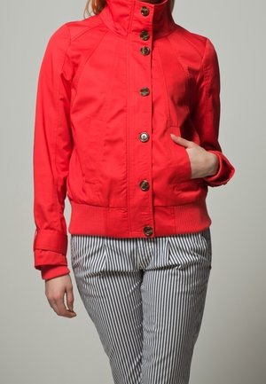 Giacca rossa attillata con collo alto, chiusura a bottoni e orlo a coste. Indossata sopra pantaloni a righe, dotati di tasche laterali e una texture liscia.