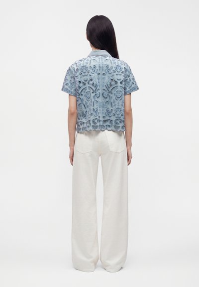 Femme de dos portant une chemise courte en dentelle bleue avec motif floral et un pantalon blanc large et ample sur fond uni.