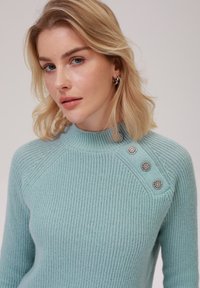 Pulover tricotat de culoare bleu deschis cu guler înalt, având trei butoane decorative argintii pe umărul stâng și un finisaj texturat.