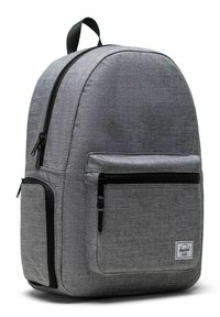Sac à dos en tissu gris de forme arrondie, zippers noirs, poche avant et un patch logo. Texture lisse avec un design minimaliste.