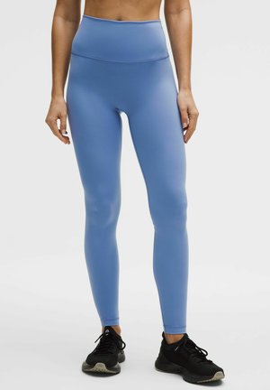 Mujer con leggings azules de tiro alto y zapatos deportivos negros, de pie contra un fondo claro y liso.