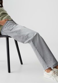 Personne portant un jean droit gris clair, des baskets blanches et un pull vert, assise de côté sur une chaise noire sur fond blanc.