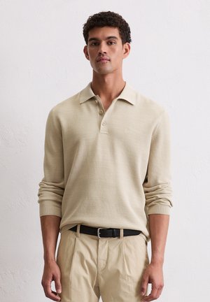 Beige Langarm-Polo mit strukturiertem Gewebe, einer Knopfleiste mit drei Knöpfen und einem weichen Kragen. Getragen mit khakifarbenen Hosen und einem schwarzen Gürtel.