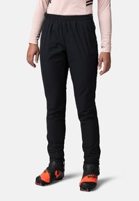 Pantalons de sport noirs avec taille élastique, en tissu léger, coupe ajustée, dotés de poches latérales et portés avec des bottes de ski à accents orange.