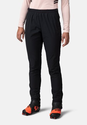 Schwarze Sporthosen mit elastischem Bund, aus leichtem Stoff, schmal zulaufende Passform, mit seitlichen Taschen und getragen mit orangefarbenen Skischuhen.