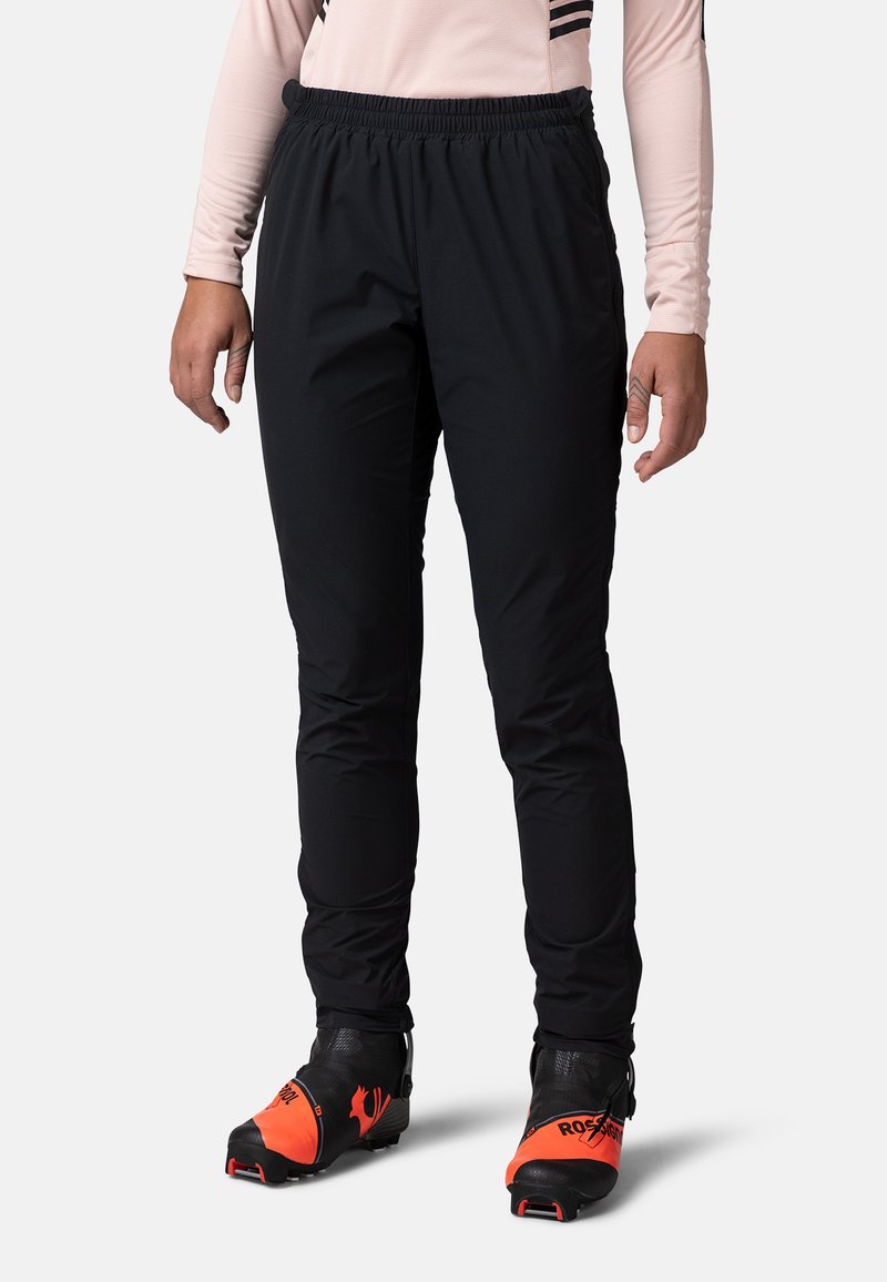 Pantaloni sportivi neri con cintura elastica, tessuto leggero, vestibilità affusolata, dotati di tasche laterali e indossati con scarponi da sci con dettagli arancioni.