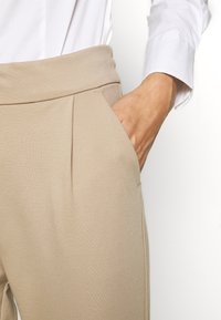 Handen i fickan på beiga högmidjade byxor som bärs med en vit långärmad skjorta på en enfärgad vit bakgrund.