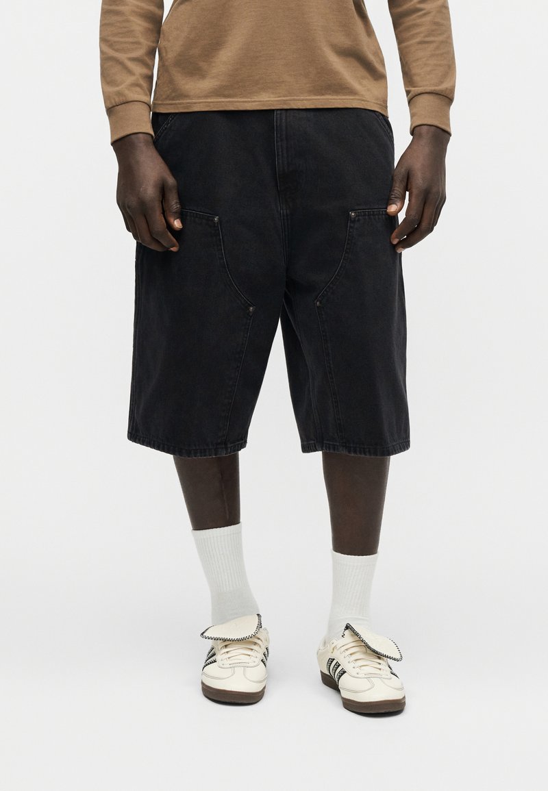 Persona che indossa pantaloncini neri fino al ginocchio, calzini bianchi e sneaker color avorio con alette sollevate, in piedi contro uno sfondo bianco semplice.