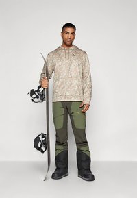 Snowboarder che indossa una felpa mimetica color sabbia e pantaloni verdi, con in mano una tavola da snowboard. Si vedono stivali neri e attacchi. Sfondo uniforme.