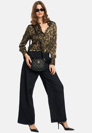 Zwarte gewatteerde schoudertas, olijfgroene blouse met patroon, wijde zwarte spijkerbroek, en zwarte puntige hakken, tegen een witte achtergrond.