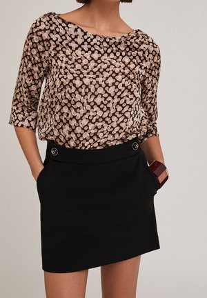 Femme portant une jupe noire avec des détails de boutons et un chemisier à manches trois-quarts avec des motifs circulaires beige et rose.