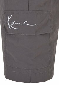 Shorts cargo gris avec une poche à rabat arborant un logo "Kari" brodé en blanc. Le tissu a une texture lisse et un léger éclat.