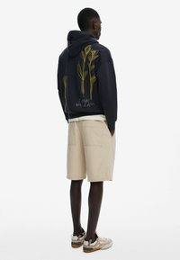 Marineblå hættebluse med gule blomsterbroderier på ryggen, parret med beige shorts og grå sneakers. Designet har hætte og lomme-detaljer.