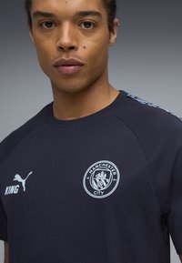 Marineblå bomulds-T-shirt med rund halsudskæring, officielt Manchester City-logo og hvide Puma-logoer. Mønsterdetalje i hvid på siden.
