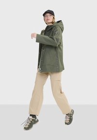 Veste imperméable vert olive avec capuche, pantalon beige et baskets vertes. Le modèle porte une casquette noire, adoptant une posture détendue en marchant.