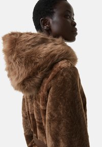 Hnědý umělý kožíšek s odnímatelnou, huňatou kapucí. Disponuje páskem v oblasti pasu a měkkou texturou, zobrazeno ze strany.