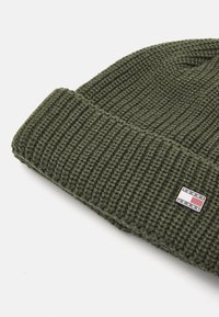 Zelena pletena beanie kapa s rebrastom teksturom i preklopnim rubom, s malim brendiranim oznakom s "TOMMY JEANS" logotipom u kontrastnim bojama.