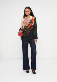 Bluză neagră cu un design floral colorat, mâneci largi; asortată cu pantaloni din denim închis și o geantă mică roșie; pantofi negri cu vârf ascuțit.