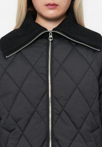 Giacca nera trapuntata con colletto a coste, cerniera argentata e motivo a diamante; il tessuto sembra liscio con una texture leggermente imbottita.