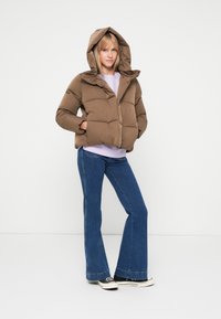 Casaco puffer castanho com capuz, apresentando um design acolchoado e fecho zip. Usado sobre um top lilás claro e combinado com jeans flare.