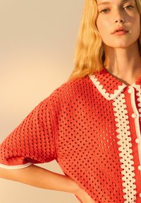 Maglietta rossa a maglia con collo bianco a scallop e chiusura con bottone; presenta una grande trama a rete e maniche corte.