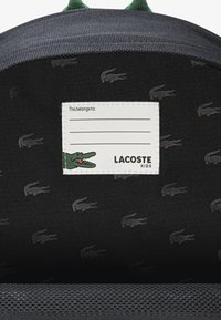 Innenraum eines schwarzen Stoffrucksacks mit wiederholtem Lacoste-Krokodilmuster. Verfügt über ein weißes Etikett mit Platz zur Personalisierung und grüne Akzente.