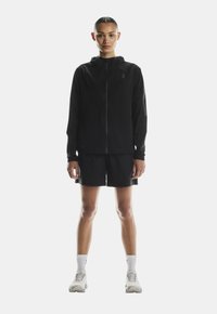 Must oruga jakk, mis on kapuutsiga ja on ühendatud mustade lühikeste pükstega. Kerge kangas, minimalistlik disain ja peegeldavad aktsendid. Sportlikud jalatsid ja sokid.