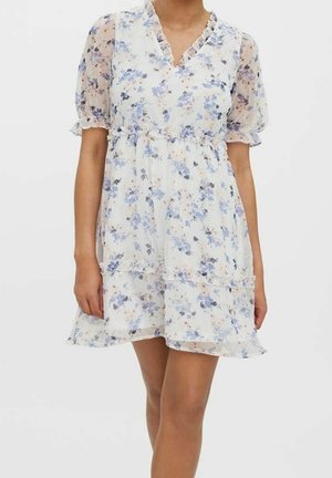 Femme portant une robe courte blanche à fleurs avec des manches bouffantes transparentes et un col en V à volants, debout devant un fond uni.