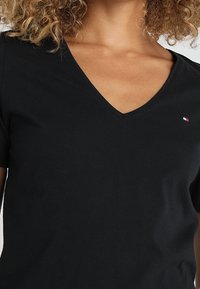 Schwarzes V-Ausschnitt-T-Shirt aus Baumwolle mit kurzen Ärmeln und einem kleinen Logoakzent in Rot, Weiß und Blau auf der linken Brust.