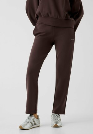 Persona che indossa pantaloni Guess marrone scuro a gamba dritta e sneakers bianche, in piedi davanti a uno sfondo chiaro e uniforme.