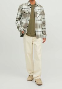 Camicia di flanella a quadri grigia e bianca, con due tasche sul petto e bottoni neri, indossata sopra una t-shirt verde oliva, abbinata a pantaloni color crema.