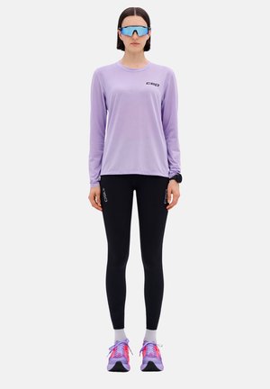 Langärmliges lila Hemd mit schwarzem Logo, schwarze Leggings und mehrfarbige Sportschuhe mit pinken Akzenten und grauen Sohlen. Blaue Sonnenbrille.