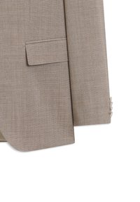 Gros plan sur la manche d'une veste de costume beige texturée avec trois boutons sur le poignet et une poche à rabat à l'avant côté gauche.