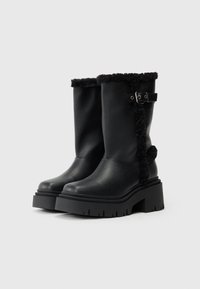 Botas de couro pretas com um punho peludo e sola espessa e plana. Apresentam uma fivela lateral e detalhes texturizados ao longo do punho.