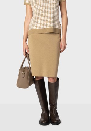 Femme portant un haut beige à motif pied-de-poule, une jupe couleur camel, des bottes hautes marron foncé, tenant un sac taupe devant un fond clair.