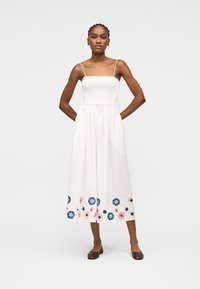 Femme debout, les mains derrière le dos, portant une robe midi blanche avec corsage smocké, fines bretelles et broderie florale à l'ourlet.