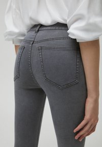 Jean en denim gris à coupe ajustée, avec deux poches arrière et une surface lisse légèrement texturée. Blouse blanche visible au-dessus.