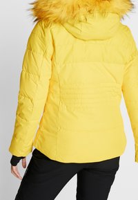 Gelbe Pufferjacke mit abnehmbarer Kunstfellkapuze, gestepptem Muster und tailliertem Schnitt, ausgestattet mit elastischen Bündchen und einem leicht abgerundeten Saum.