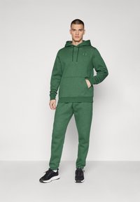 Felpa verde con cappuccio e pantaloni jogger coordinati realizzati in tessuto morbido. La felpa presenta una tasca a marsupio e un cordoncino regolabile. Sneakers nere.